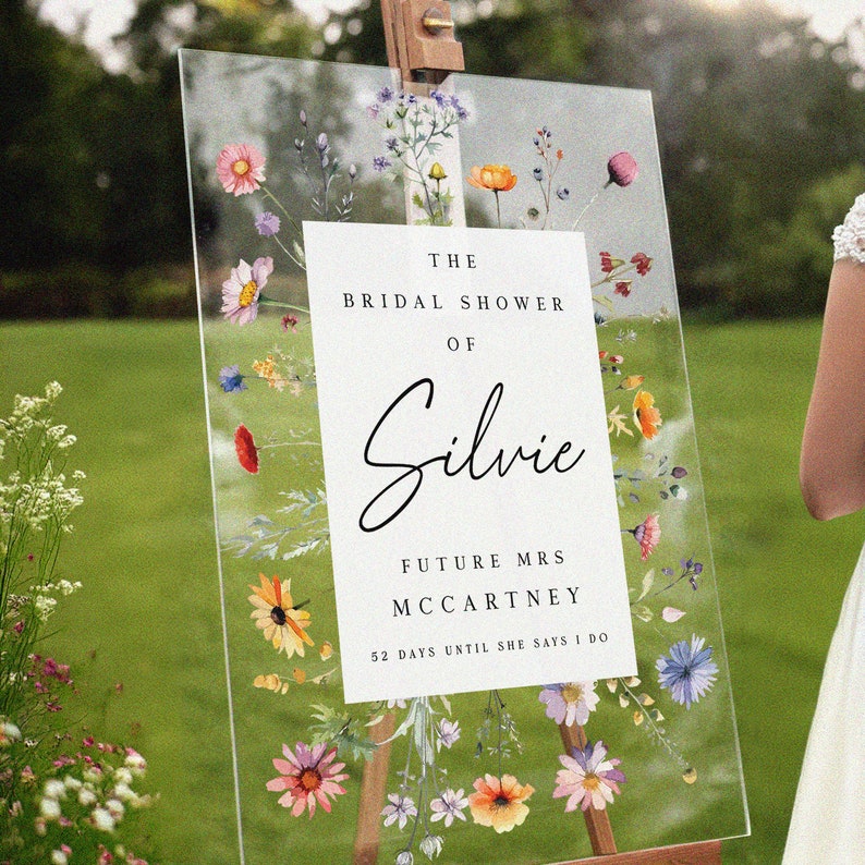 May include: A clear acrylic rectangular sign featuring a white center panel with the text THE BRIDAL SHOWER OF Silvie FUTURE MRS MCCARTNEY 52 DAYS UNTIL SHE SAYS I DO, surrounded by a colorful watercolor floral border of wildflowers and greenery.
