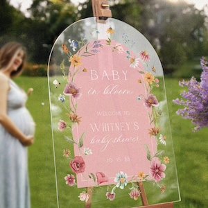 Op de afbeelding: Een helder acrylbord voor een babyshower met een roze midden en een bloemenrand. Op het bord staat "BABY in bloom" en verwelkomt gasten bij "Whitney's babyshower" met een datum. Het bord staat op een houten ezel.