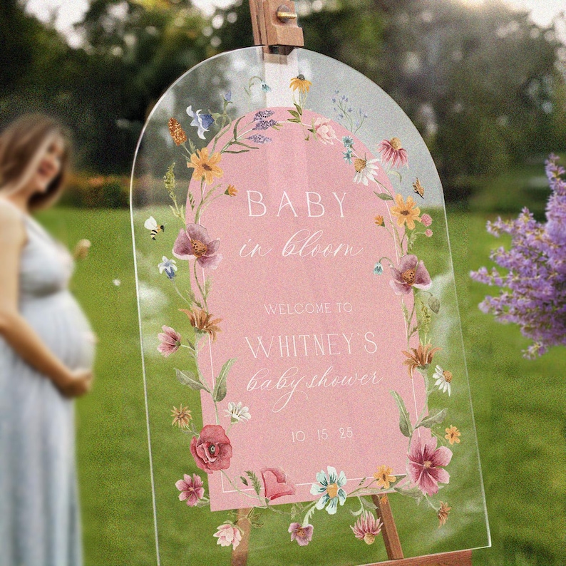 Op de afbeelding: Een helder acrylbord voor een babyshower met een roze midden en een bloemenrand. Op het bord staat "BABY in bloom" en verwelkomt gasten bij "Whitney's babyshower" met een datum. Het bord staat op een houten ezel.