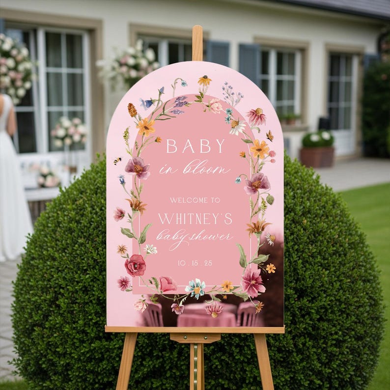 Op de afbeelding: Een roze welkomstbord op een houten ezel, met de tekst "BABY in bloom" en "WHITNEY'S babyshower". Het bord is versierd met bloemenillustraties in roze, gele en paarse tinten. De datum is 15 oktober 25.