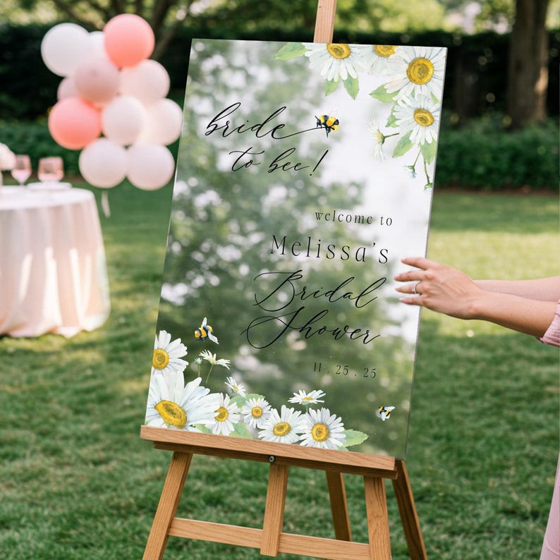 May include: A clear acrylic sign on a wooden easel with the text "Bride to bee!" and "Melissa's Bridal Shower" with a date. The sign is decorated with watercolor daisies and bees. The sign is displayed outdoors.