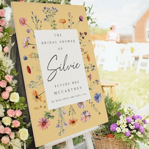 May include: A rectangular sign with a mustard yellow background featuring a white central panel. The text reads THE BRIDAL SHOWER OF Silvie FUTURE MRS MCCARTNEY 52 DAYS UNTIL SHE SAYS I DO. The sign is decorated with watercolor wildflowers.