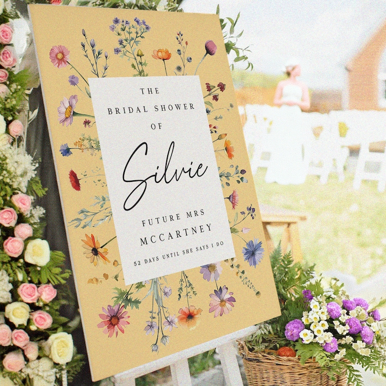 May include: A rectangular sign with a mustard yellow background featuring a white central panel. The text reads THE BRIDAL SHOWER OF Silvie FUTURE MRS MCCARTNEY 52 DAYS UNTIL SHE SAYS I DO. The sign is decorated with watercolor wildflowers.