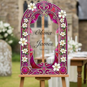 May include: An arched wedding sign featuring a stained glass pattern in shades of magenta and red with white floral accents. The text reads: WELCOME TO Olivia & James WEDDING 03.04.26. The sign is mounted on a wooden easel stand.