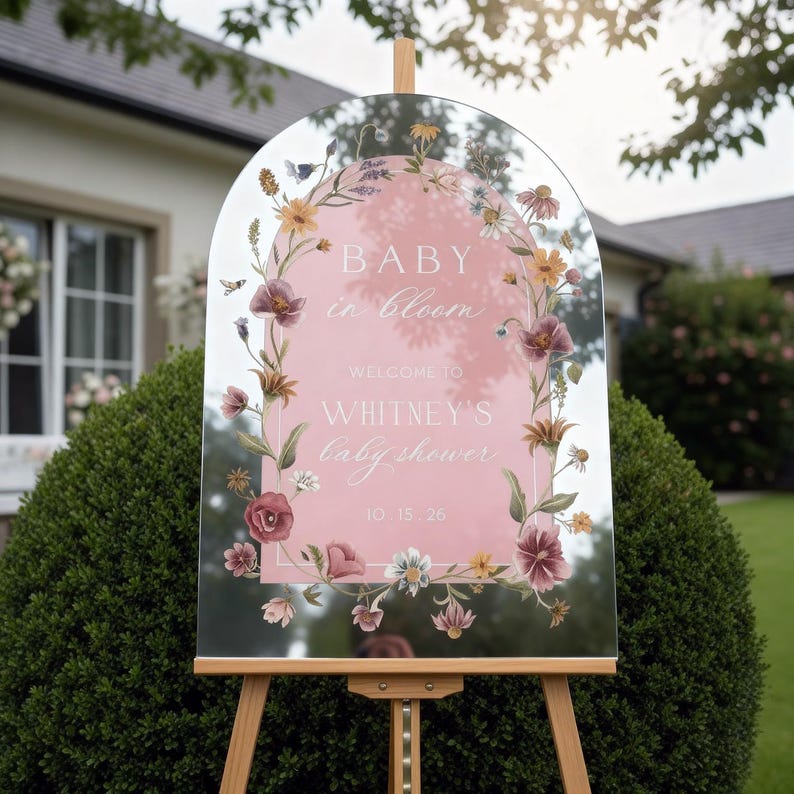 Op de afbeelding: Een helder, boogvormig bord op een houten ezel, met een roze middenstuk met de tekst "BABY in bloom" en "WELCOME TO WHITNEY'S baby shower", omgeven door een bloemenontwerp. De datum "10.15.26" staat onderaan.