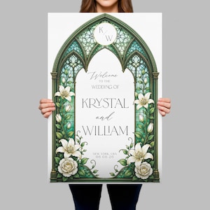 K&ouml;nnte beinhalten: Ein rechteckiges Hochzeits-Willkommensschild mit einem gr&uuml;nen Buntglas-Bogen-Design, verziert mit wei&szlig;en Lilien und Rosen. Der Text lautet: K/W, Welcome to the wedding of Krystal and William, New York, USA, 06.06.26. Das Schild hat einen wei&szlig;en Hintergrund.