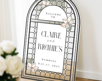 May include: An arched wedding welcome sign featuring a black stained-glass window frame design. The text reads: WELCOME TO CLAIRE and RICHIE'S WEDDING MAY 17, 2025. The sign is decorated with pale pink magnolia flowers and green leaves.