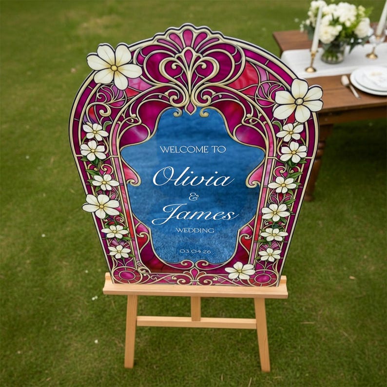 May include: A wedding welcome sign featuring a stained glass style border in shades of magenta and deep pink with white floral accents. The centre is blue with white text reading Welcome to Olivia & James Wedding 03.04.26 on a wooden easel.