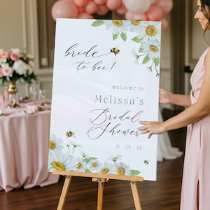 May include: A white sign on a wooden easel with the text "Bride to bee!" and "Melissa's Bridal Shower" in elegant script. The sign is decorated with daisies and bees, with the date 11.25.25.