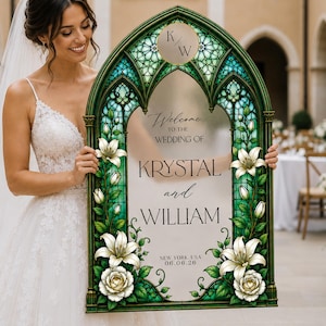 K&ouml;nnte beinhalten: Ein bogenf&ouml;rmiges Hochzeits-Willkommensschild mit einem gr&uuml;nen Buntglasrahmen und wei&szlig;en Lilien- sowie Rosenmotiven. Das mittlere Paneel zeigt den Text: Welcome to the wedding of Krystal and William, New York, USA, 06.06.26, sowie ein K/W-Monogramm.