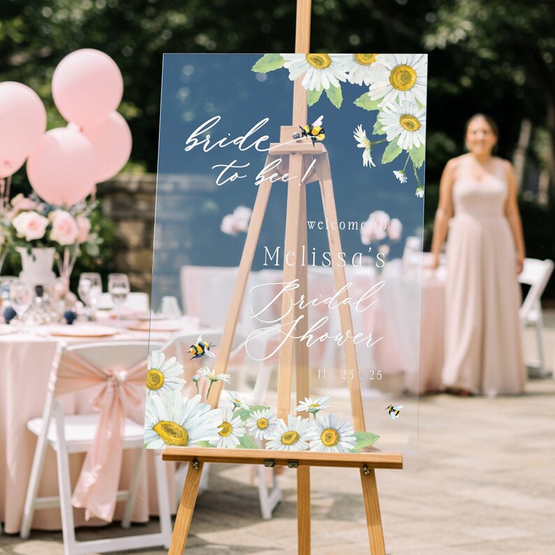 May include: A clear acrylic bridal shower welcome sign on a wooden easel. The sign features the text "Bride to bee!" and "Melissa's Bridal Shower" with daisy and bee illustrations. The date "11.25.25" is also displayed.