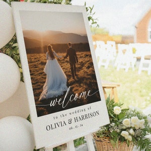 May include: A white wedding welcome sign with a photo of a couple walking in a field at sunset. The sign says "Welcome to the wedding of Olivia & Harrison 06.15.24".