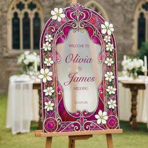 May include: An arched wedding welcome sign featuring a stained glass pattern in shades of magenta and deep red, decorated with white flowers. The text reads: WELCOME TO Olivia & James WEDDING 03.04.26. The sign is mounted on a wooden easel.