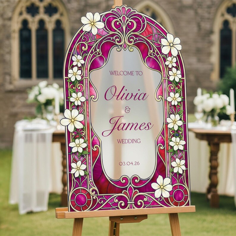 May include: An arched wedding welcome sign featuring a stained glass pattern in shades of magenta and deep red, decorated with white flowers. The text reads: WELCOME TO Olivia & James WEDDING 03.04.26. The sign is mounted on a wooden easel.