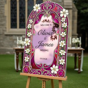 May include: An arched wedding welcome sign featuring a stained glass design in shades of magenta and gold, adorned with white flowers. The text reads: WELCOME TO Olivia & James WEDDING 03.04.26. The sign is displayed on a wooden easel.
