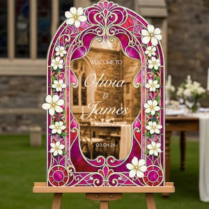 May include: An arched wedding welcome sign featuring a vibrant magenta and purple stained glass pattern with white floral accents. The centre text reads: WELCOME TO Olivia & James WEDDING 03.04.26. The sign is displayed on a wooden easel stand.