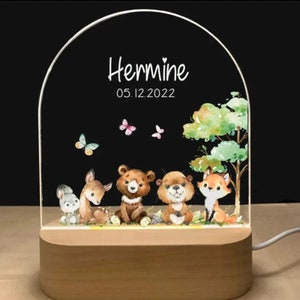 May include: A white acrylic night light with a wooden base. The light features a woodland scene with a deer, bear, beaver, and fox. The light is personalized with the name "Hermine" and the date "05.12.2022".