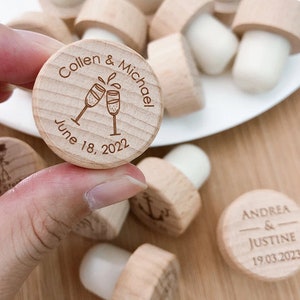 Può includere: Tappi per bottiglie in legno con testo inciso personalizzato. I tappi presentano un design con due bicchieri di champagne con una data e nomi. Il testo su un tappo dice "Colleen & Michael June 18, 2022". Il testo su un altro tappo dice "Andrea & Justine 19.03.2023".