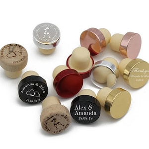 Könnte beinhalten: Sortiment an personalisierten Flaschenverschlüssen in verschiedenen Farben, darunter Holz, Schwarz, Gold, Silber und Rot. Einige Verschlüsse sind mit gravierten Namen und Daten versehen, wie z. B. "Amanda & Alex 13.07.2018" und "Alex & Amanda 18.08.18".