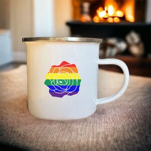 Puede incluir: Taza de esmalte blanco con un diseño de rosa arcoíris. La taza tiene un borde y un asa plateados.