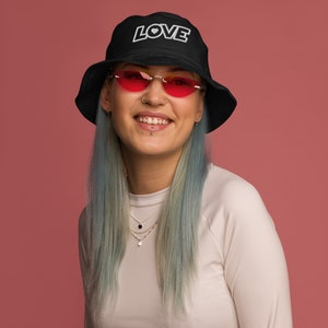 May include: A black bucket hat with white embroidery that says "LOVE" with a small heart in the "O". The model is wearing the hat and has blue hair and pink heart-shaped sunglasses.