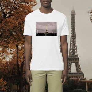 Può includere: Una maglietta bianca con una foto della Torre Eiffel a Parigi, Francia. La foto mostra la torre illuminata di notte con un cielo nuvoloso sullo sfondo.
