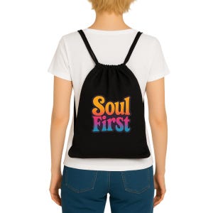 Könnte beinhalten: Schwarzer Kordelzugrucksack mit dem Text "Soul First" in einem Farbverlauf aus Orange, Pink und Blau. Die Tasche hat schwarze Träger und wird über einem weißen T-Shirt und einer blauen Jeans getragen.