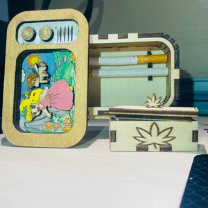 May include: A wooden box shaped like a vintage television set with a colorful cartoon scene on the front, featuring a smaller rectangular wooden box with a laser-cut leaf design on the lid, and two cigarettes resting inside the open main compartment.