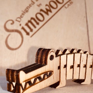 May include: A small wooden crocodile figurine constructed from layered light-colored wood with dark laser-cut edges. The crocodile features a jagged back, a wide open mouth with visible teeth, and a circular eye. The text "Designed and Made by Simowood LAB" is visible in the background.
