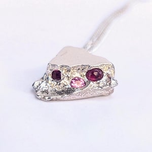 Puede incluir: Colgante plateado con superficie texturizada, con tres piedras engastadas: dos rojas oscuras y una rosa. El colgante está sujeto a un cordón blanco, sobre un fondo blanco.