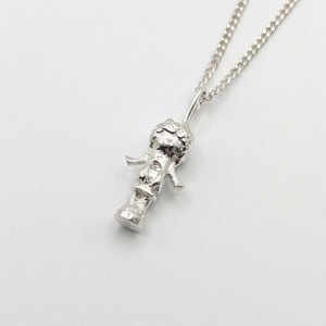 May include: A silver-toned metal pendant shaped like a small figure with a rounded head and outstretched arms, suspended from a delicate matching silver-toned chain. The pendant features a textured surface and a cylindrical base, creating a detailed charm.