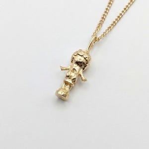 May include: A gold-coloured necklace featuring a small, three-dimensional pendant shaped like a stylised figure with a rounded head, outstretched arms, and a cylindrical base, suspended from a delicate, fine-linked gold-coloured metal chain.