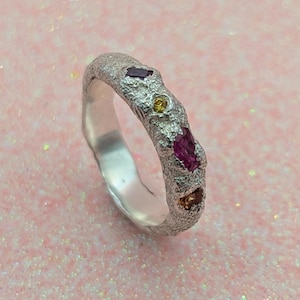Könnte beinhalten: Ein silberner Ring mit einer strukturierten Band und vier kleinen Edelsteinen. Die Edelsteine sind rosa, gelb und orange.
