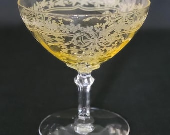 Pode incluir: Uma taça de champanhe estilo coupe com um tom amarelo. A borda da taça apresenta um desenho floral gravado. A haste e a base são transparentes. A taça é provavelmente feita de cristal ou material semelhante.