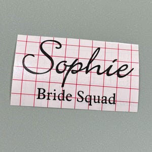 May include: A white sticker with black text that says "Sophie Bride Squad" on a red and white grid background.