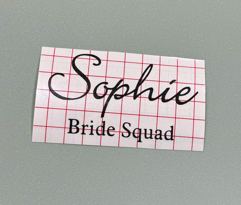 May include: A white sticker with black text that says "Sophie Bride Squad" on a red and white grid background.