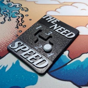 May include: A black plastic gear shift knob with white lettering that reads "THE NEED SPEED" and a white button.