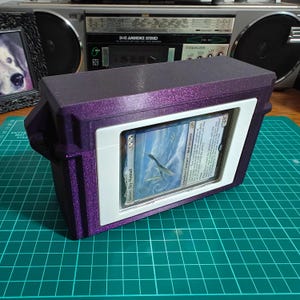 May include: A rectangular purple glitter storage box featuring a white frame on the front that holds a Magic: The Gathering trading card titled Yorion, Sky Nomad. The card displays a blue and white sky illustration with text and a 4/5 power rating.