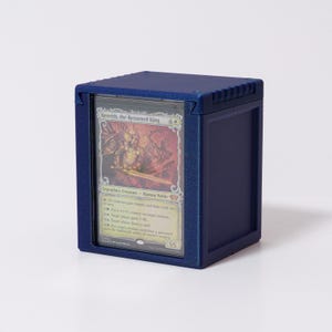 May include: A navy blue card storage box with a clear window displaying a Magic: The Gathering card. The card features an illustration of a character named "Kenrith, the Returned King". The box is square and has a textured surface.