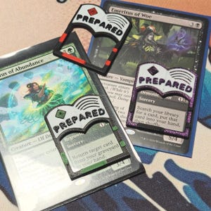 Puede incluir: Tres marcadores de cartas de plástico impresos en 3D para juegos de mesa, con forma de libro abierto y la palabra PREPARED en relieve. Un marcador es negro con detalles rojos, otro negro con verdes y otro negro con purpurina morada.