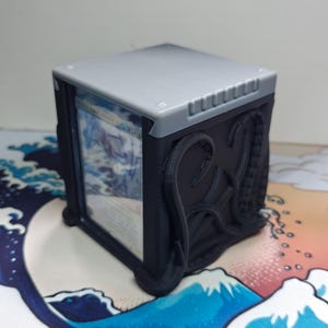 May include: A black and gray card storage box with an intricate tentacle design on the side. The box has a clear front panel displaying a card. The top of the box is gray.