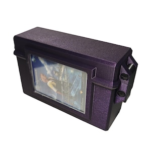 May include: A rectangular, dark purple storage box with a textured finish, featuring a clear display window on the front side. A single trading card is visible inside the window, showing a character illustration and printed game text.