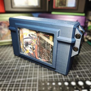 May include: A matte blue 3D-printed deck box featuring a rectangular display window on the front. A trading card titled Pymark, the Inspiration is visible inside the case. The box has a textured surface and is secured with two black screws on the side.
