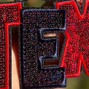 May include: Close-up of a sequined patch spelling out the word "TEXAS". The letters are outlined in black, with the "T" and "X" in red sequins and the "E" and "A" in blue sequins.