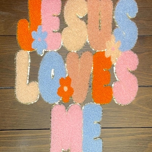 May include: A fuzzy felt applique with the words "Jesus Loves Me" in a retro style. The letters are in shades of pink, orange, blue, and beige with gold trim. Each letter has a small flower design.