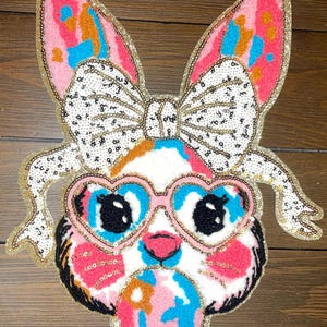 May include: A pink and blue sequined bunny rabbit with a white bow, heart-shaped sunglasses, and a bubblegum-like object in its mouth.