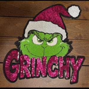 May include: A green Grinch face with a Santa hat and the word "GRINCHY" in pink sequins. The Grinch has an angry expression with green eyes and a stitched texture. The hat is pink with a white trim.
