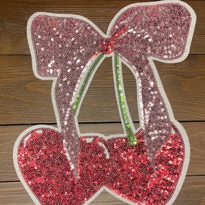 May include: A pink and red sequined cherry with a green stem and a pink bow. The cherry is on a white background.