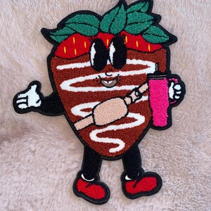 May include: A chocolate-covered strawberry cartoon character with white icing, wearing red shoes and holding a pink drink. The character has large eyes, a smile, and a straw in its mouth.