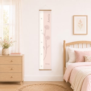 May include: A vertical wall-mounted growth chart featuring a light pink background with a white side panel containing black measurement markings. The chart displays a line-art daisy illustration, the name Amelia, and the word Daisy in dark text.